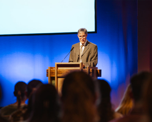 Prof. Dr. Markus Zehnder steht allein hinter einem hölzernen Rednerpult auf einer Bühne. Er trägt einen hellbraunen Anzug mit einem weißen Hemd und einer beigefarbenen Krawatte. Der Mann hat graue Haare und trägt eine Brille. Er schaut konzentriert auf seine Unterlagen oder in die Richtung des Publikums. Im unteren Bildbereich sind die unscharfen Silhouetten von Zuhörern zu sehen, die aufmerksam in Richtung des Redners blicken. Die Atmosphäre wirkt ruhig, konzentriert und würdevoll – passend für eine akademische Veranstaltung.