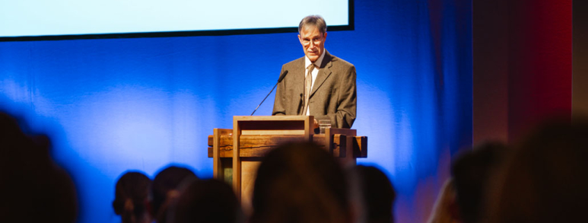 Prof. Dr. Markus Zehnder steht allein hinter einem hölzernen Rednerpult auf einer Bühne. Er trägt einen hellbraunen Anzug mit einem weißen Hemd und einer beigefarbenen Krawatte. Der Mann hat graue Haare und trägt eine Brille. Er schaut konzentriert auf seine Unterlagen oder in die Richtung des Publikums. Im unteren Bildbereich sind die unscharfen Silhouetten von Zuhörern zu sehen, die aufmerksam in Richtung des Redners blicken. Die Atmosphäre wirkt ruhig, konzentriert und würdevoll – passend für eine akademische Veranstaltung.