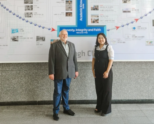 Dr. Friedemann Burkhardt steht gemeinsam mit einer Frau vor einer Wandgrafik an der Handong Global University in Südkorea. Auf der Grafik sind die Worte ‚Honesty, Integrity and Faith‘ zu lesen