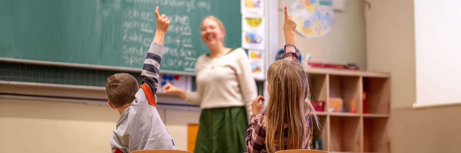 Zwei Kinder sitzen in einem Klassenzimmer und melden sich, während eine Lehrerin vor der Tafel unterrichtet.
