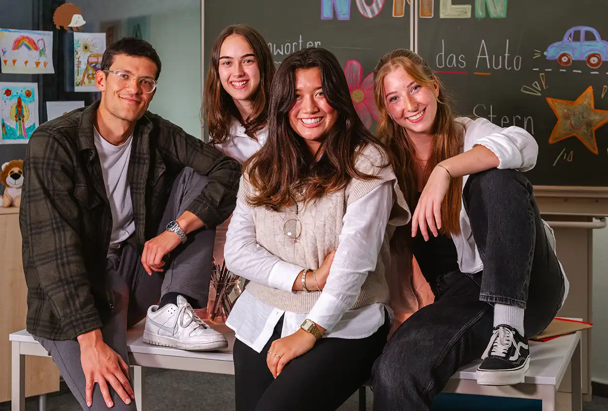 Drei Studentinnen und ein Student sitzen in lockerer Haltung auf einem Tisch in einem Grundschulklassenzimmer. Im Hintergrund ist eine mit Kreide beschriebene grüne Tafel zu sehen.