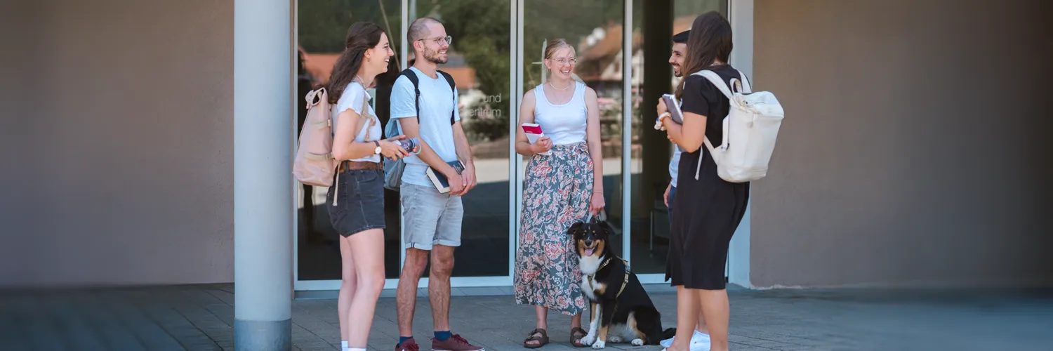 Zwei Studenten und drei Studentinnen stehen mit einem angeleinten Hund vor der Haupteingangstür der IHL.