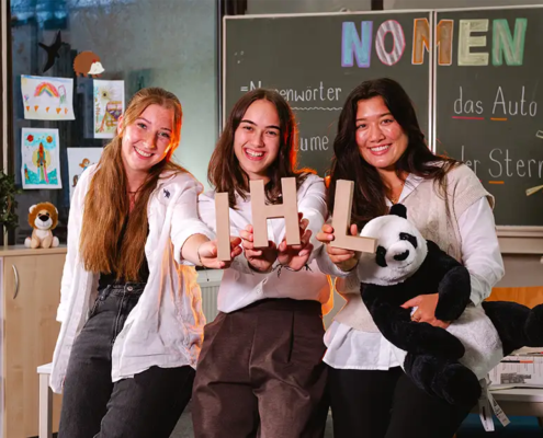 3 Studentinnen sitzen lächelnd im Klassenzimmer einer Grundschule und halten die Buchstaben IHL in die Kamera