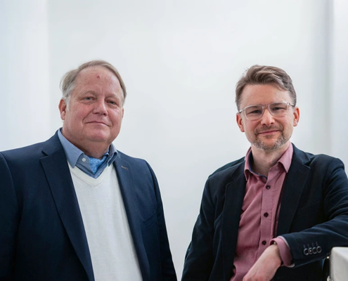Daniel Strass und Arndt Schnepper stehen zusammen in einem Lichtdurchfluteten Gebäude.