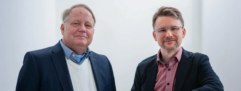 Daniel Strass und Arndt Schnepper stehen zusammen in einem Lichtdurchfluteten Gebäude.