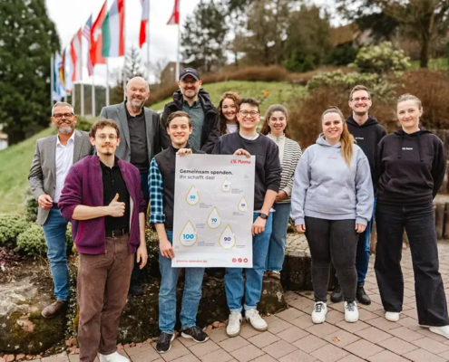„Eine Gruppe Studierender steht gemeinsam mit Vertretern der Hochschule, der Liebenzeller Mission und Mitarbeitenden von CSL Plasma für ein Gruppenfoto. Sie halten ein Plakat mit der Aufschrift ‚Gemeinsam spenden. Ihr schafft das.‘.“