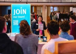 Im Vordergrund sind mehrere Studierende zu sehen, die einer Referentin bei der Berufsmesse zuhören. Im Hintergrund sieht man ebenfalls viele Studierende, die an anderen Stellen anderen Referenten zuhören.