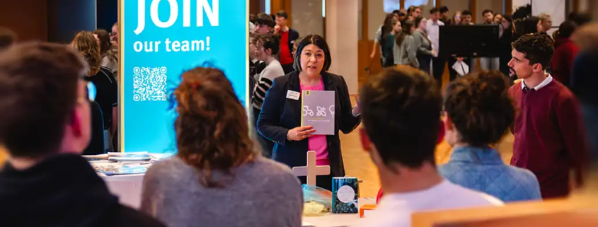 Im Vordergrund sind mehrere Studierende zu sehen, die einer Referentin bei der Berufsmesse zuhören. Im Hintergrund sieht man ebenfalls viele Studierende, die an anderen Stellen anderen Referenten zuhören.