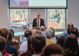 Professor David Kramer hält seine Antrittsvorlesung in den Räumlichkeiten der Internationalen Hochschule Liebenzell. Das Bild zeigt ihn an einem hellen Sommerabend vor einem großen, sitzenden Publikum im vollen Vortragssaal der IHL.