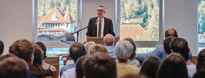 Professor David Kramer hält seine Antrittsvorlesung in den Räumlichkeiten der Internationalen Hochschule Liebenzell. Das Bild zeigt ihn an einem hellen Sommerabend vor einem großen, sitzenden Publikum im vollen Vortragssaal der IHL.