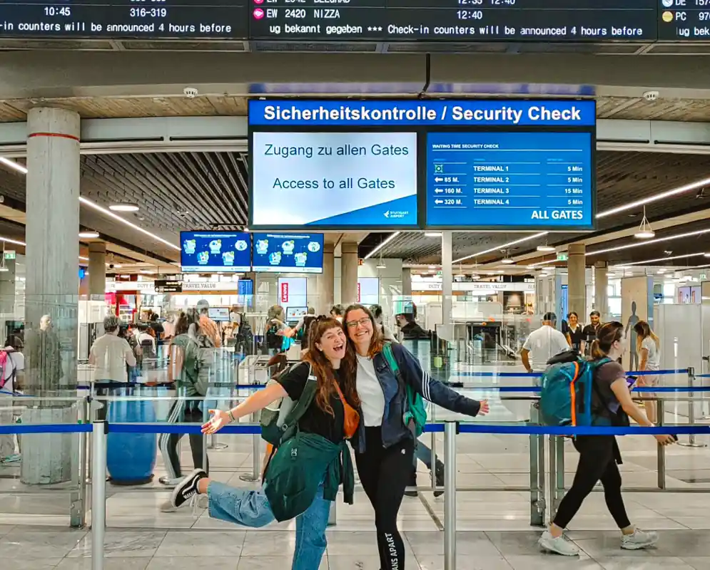 Zwei freudestrahlende Studentinnen der Internationalen Hochschule Liebenzell stehen mit Rucksäcken vor der Sicherheitskontrolle am Flughafen Stuttgart. Sie posieren voller Vorfreude vor der Anzeigetafel zum Abflug ins Auslandssemester.
