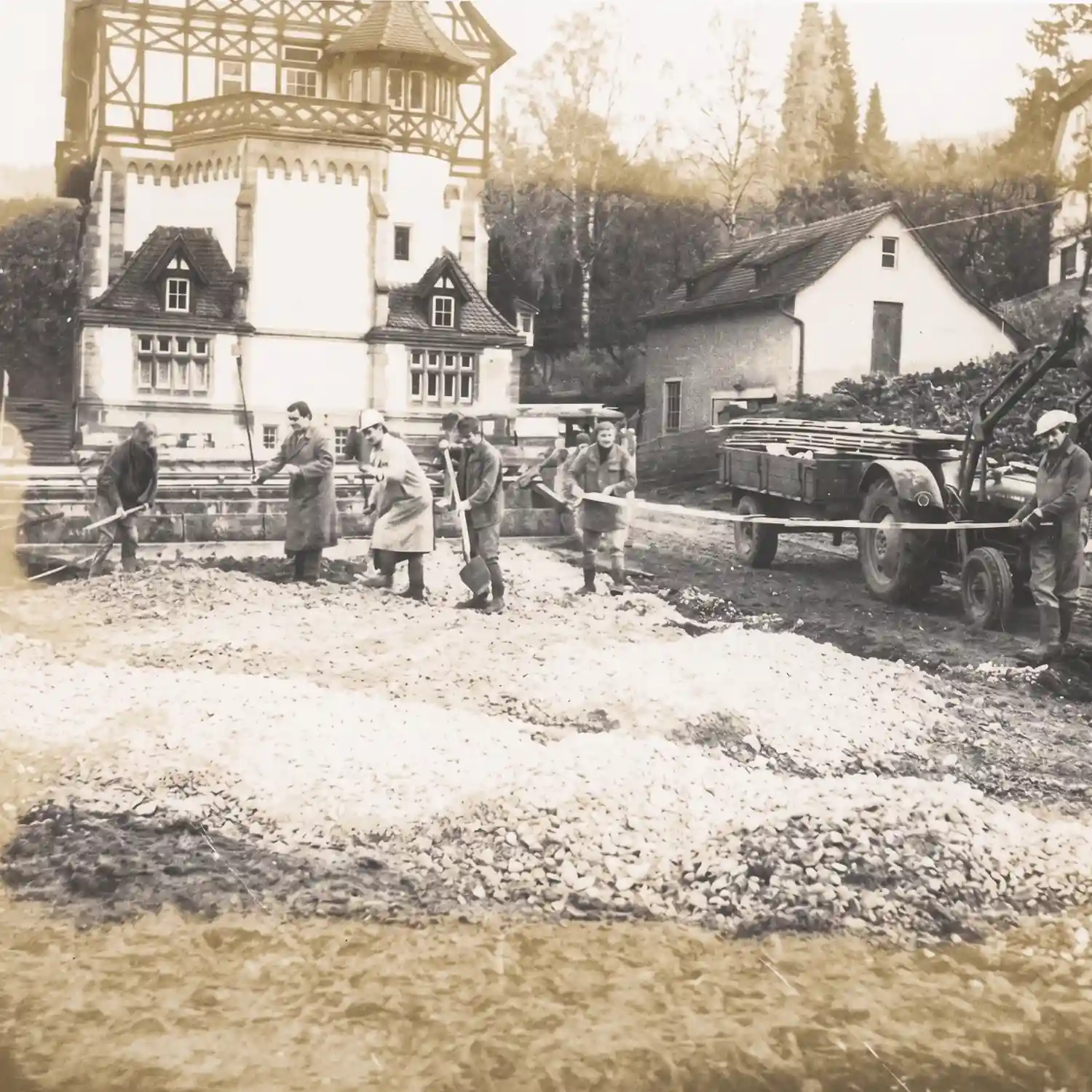 Eine historische Schwarz-Weiß-Aufnahme zeigt Bauarbeiten auf dem Gelände der Liebenzeller Mission in Bad Liebenzell. Mehrere Männer arbeiten mit Schaufeln an Kieshaufen und einem Traktor mit Anhänger, während im Hintergrund das historische Fachwerk-Missionshaus zu sehen ist.