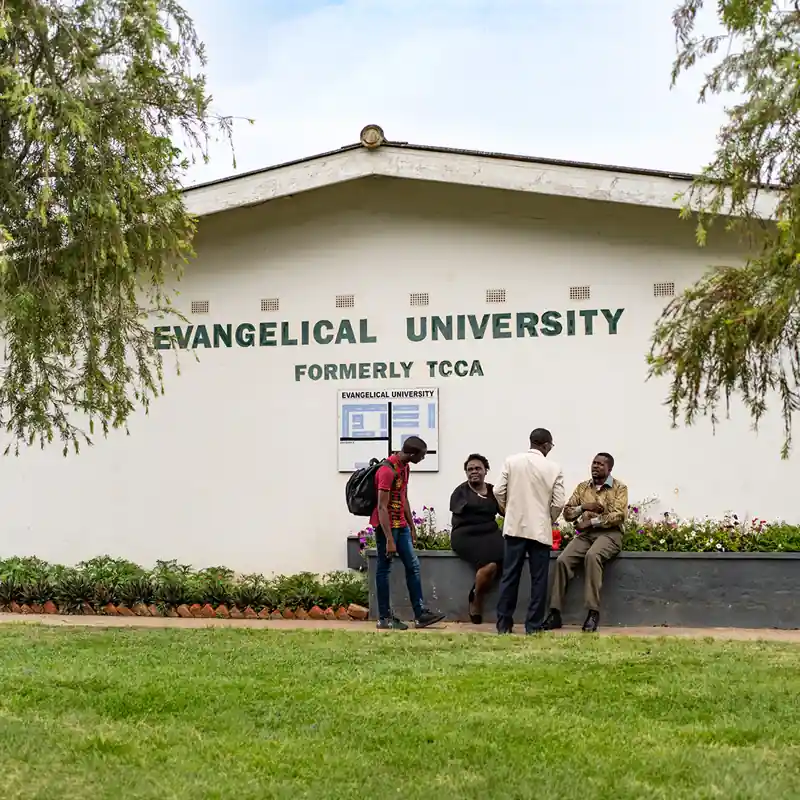 Aussenansicht der Evangelical University in Sambia (ehemals TCCA), einer Partnerhochschule der Internationalen Hochschule Liebenzell. Vor der weissen Gebaeudewand mit dem Hochschulschriftzug stehen und sitzen Studierende sowie Dozenten im Gespraech.