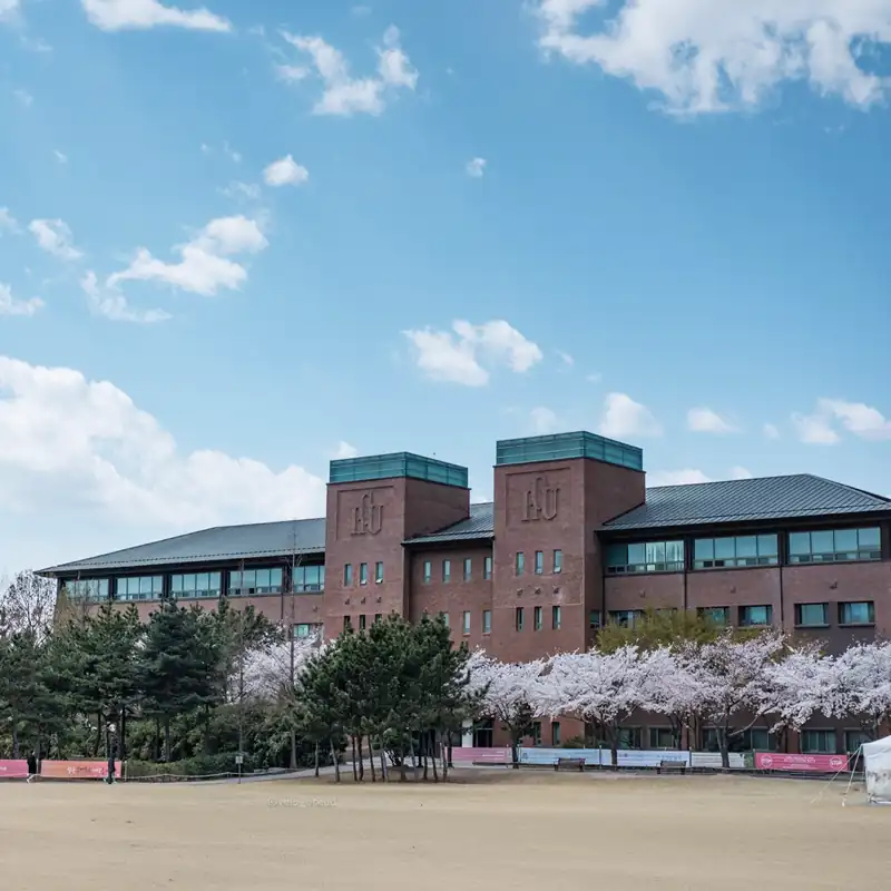 Der Campus der Handong Global University (HGU) in Südkorea, einer Partnerhochschule der Internationalen Hochschule Liebenzell. Zu sehen ist ein imposantes rotes Backsteingebäude mit HGU-Logo unter strahlend blauem Himmel, davor stehen blühende Kirschbäume und grüne Nadelbäume am Rande einer freien Fläche.