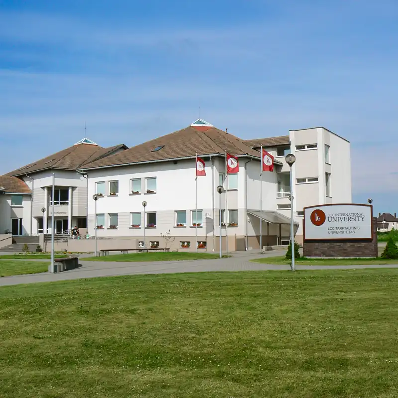 Der Campus der LCC International University in Klaipėda, Litauen, einer Partnerhochschule der Internationalen Hochschule Liebenzell. Das Bild zeigt das moderne Hochschulgebäude bei sonnigem Wetter mit roter Beflaggung, einer gepflegten Grünfläche im Vordergrund und einem großen Eingangsschild der Universität.