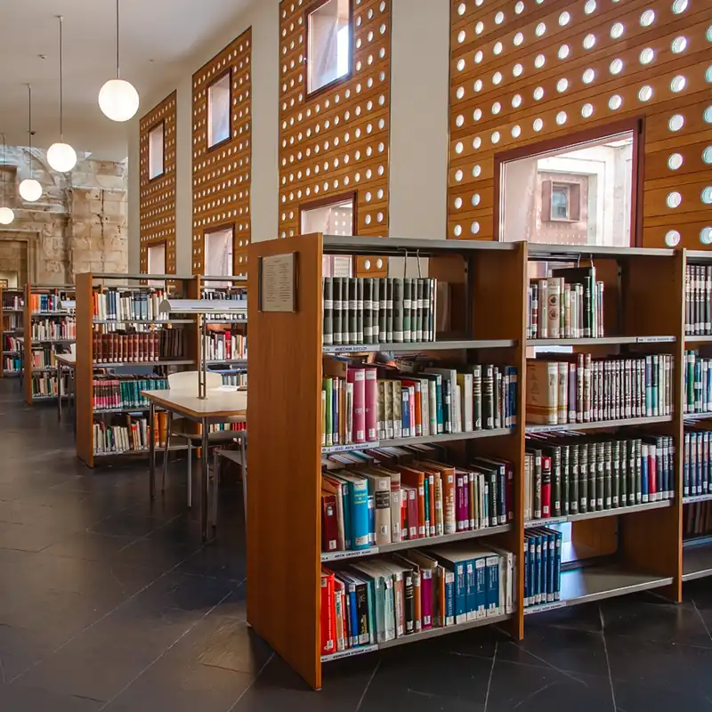 Die Bibliothek der Pontifical University of Salamanca in Spanien, einer Partnerhochschule der Internationalen Hochschule Liebenzell. Zu sehen sind gut gefüllte Bücherregale aus Holz, Lernplätze für Studierende und eine charakteristische Wandgestaltung mit Holzverkleidung, runden Glaselementen und Hängeleuchten.