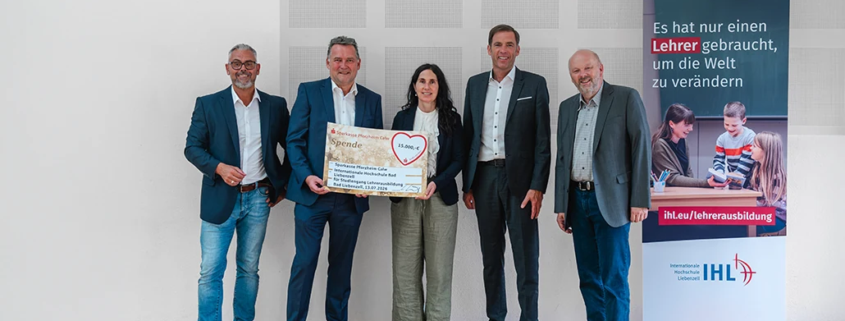 Feierliche Übergabe einer Spende in Höhe von 15.000 Euro der Sparkasse Pforzheim Calw für den Studiengang Lehrerausbildung an der Internationalen Hochschule Liebenzell. Von links nach rechts stehen Thomas Haid (Kaufmännischer Geschäftsführer der Liebenzeller Mission), der Schömberger Filialdirektor Jens Teufel, Hanna Reuther (Kanzlerin der IHL), Sven Eisele (Vorsitzender des Vorstands der Sparkasse Pforzheim Calw) und Rektor Prof. Dr. Volker Gäckle auf Holzboden vor einer weißen Wand. Sie halten gemeinsam den großen Spendenscheck. Rechts im Bild steht ein Roll-Up-Banner der IHL mit der Aufschrift: Es hat nur einen Lehrer gebraucht, um die Welt zu verändern.