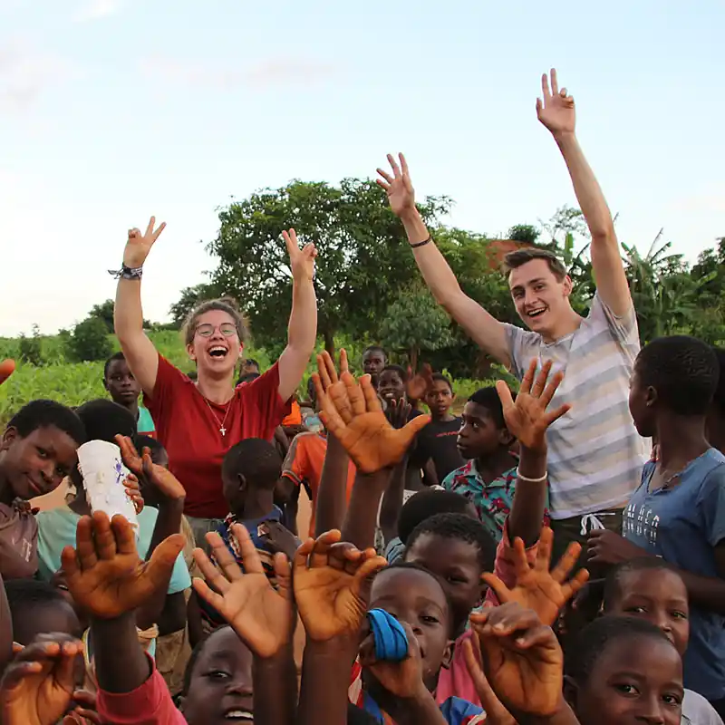 Zwei Studierende der Internationalen Hochschule Liebenzell stehen freudestrahlend mit erhobenen Händen inmitten einer Gruppe von fröhlichen Kindern in Malawi während ihres Missionseinsatzes.
