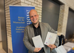 Dr. habil. Andreas Heidel, Dozent an der Internationalen Hochschule Liebenzell, steht lächelnd vor dem Messestand des Mohr Siebeck Verlags auf der Tagung der Studiorum Novi Testamenti Societas (SNTS) in Durham. Er hält seine beiden Fachpublikationen „Religiöse Erfahrung in Korinth“ und „Early Christian Centers: Corinth“ in den Händen.