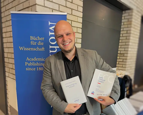 Dr. habil. Andreas Heidel, Dozent an der Internationalen Hochschule Liebenzell, steht lächelnd vor dem Messestand des Mohr Siebeck Verlags auf der Tagung der Studiorum Novi Testamenti Societas (SNTS) in Durham. Er hält seine beiden Fachpublikationen „Religiöse Erfahrung in Korinth“ und „Early Christian Centers: Corinth“ in den Händen.