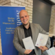 Dr. habil. Andreas Heidel, Dozent an der Internationalen Hochschule Liebenzell, steht lächelnd vor dem Messestand des Mohr Siebeck Verlags auf der Tagung der Studiorum Novi Testamenti Societas (SNTS) in Durham. Er hält seine beiden Fachpublikationen „Religiöse Erfahrung in Korinth“ und „Early Christian Centers: Corinth“ in den Händen.