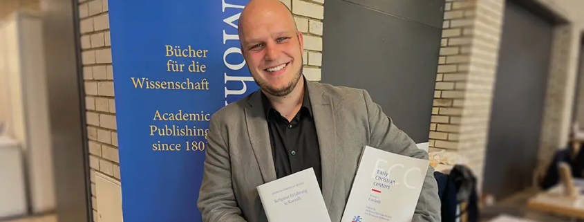 Dr. habil. Andreas Heidel, Dozent an der Internationalen Hochschule Liebenzell, steht lächelnd vor dem Messestand des Mohr Siebeck Verlags auf der Tagung der Studiorum Novi Testamenti Societas (SNTS) in Durham. Er hält seine beiden Fachpublikationen „Religiöse Erfahrung in Korinth“ und „Early Christian Centers: Corinth“ in den Händen.