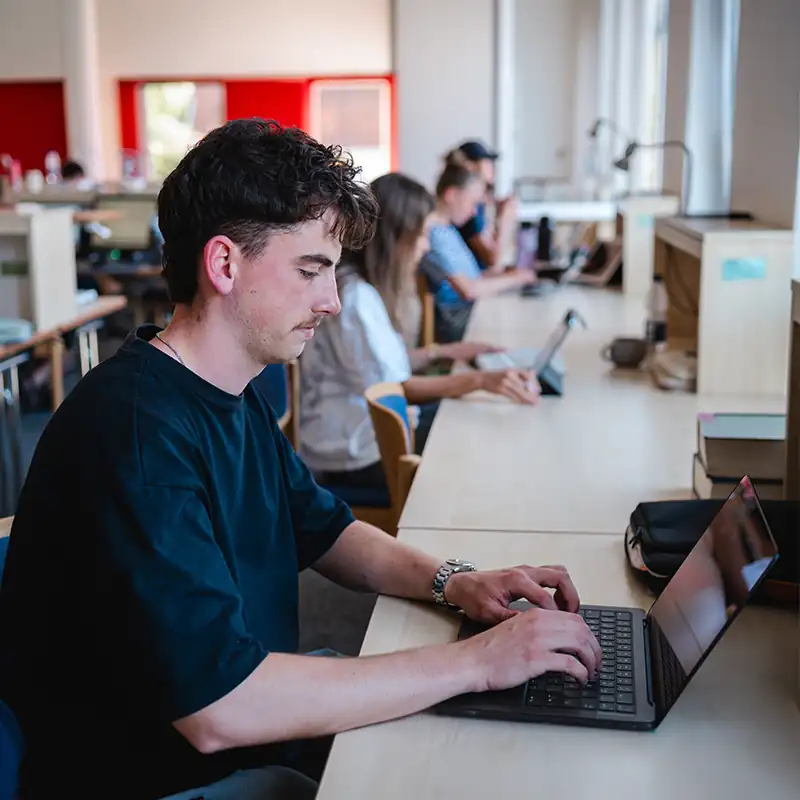 Ein Student im dunklen T-Shirt sitzt konzentriert an einer langen Tischreihe in der Bibliothek der Internationalen Hochschule Liebenzell und tippt auf seinem Laptop. Im Hintergrund arbeiten weitere Studierende an ihren Arbeitsplätzen.