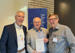 Prof. Roland Deines überreicht Prof. Karl-Wilhelm Niebuhr im Beisein von Jens Herzer am Stand des Mohr Siebeck Verlags das erste Exemplar des neuen CJHNT-Tagungsbands. Die drei Wissenschaftler stehen lächelnd nebeneinander, während Prof. Niebuhr das Buch in den Händen hält.