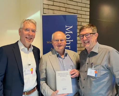 Prof. Roland Deines überreicht Prof. Karl-Wilhelm Niebuhr im Beisein von Jens Herzer am Stand des Mohr Siebeck Verlags das erste Exemplar des neuen CJHNT-Tagungsbands. Die drei Wissenschaftler stehen lächelnd nebeneinander, während Prof. Niebuhr das Buch in den Händen hält.