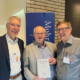 Prof. Roland Deines überreicht Prof. Karl-Wilhelm Niebuhr im Beisein von Jens Herzer am Stand des Mohr Siebeck Verlags das erste Exemplar des neuen CJHNT-Tagungsbands. Die drei Wissenschaftler stehen lächelnd nebeneinander, während Prof. Niebuhr das Buch in den Händen hält.