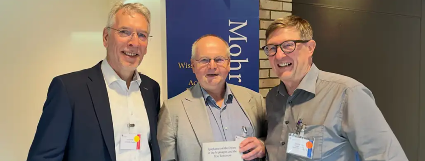 Prof. Roland Deines überreicht Prof. Karl-Wilhelm Niebuhr im Beisein von Jens Herzer am Stand des Mohr Siebeck Verlags das erste Exemplar des neuen CJHNT-Tagungsbands. Die drei Wissenschaftler stehen lächelnd nebeneinander, während Prof. Niebuhr das Buch in den Händen hält.