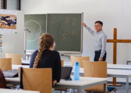 Prof. Dr. Shin Yoshida von der Tohoku Gakuin Universität lehrt während der September School 2026 an der Internationalen Hochschule Liebenzell. Er steht lächelnd vor einer Wandtafel mit japanischen Schriftzeichen und zeigt auf die Tafel, während Studierende im Seminarraum zuhören.