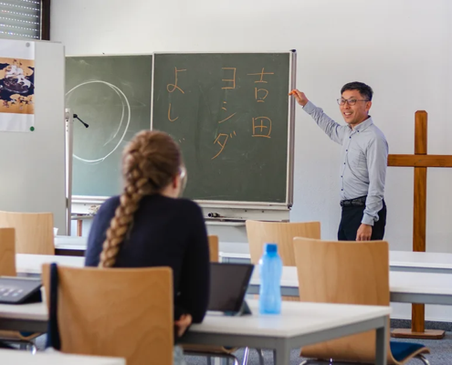 Prof. Dr. Shin Yoshida von der Tohoku Gakuin Universität lehrt während der September School 2026 an der Internationalen Hochschule Liebenzell. Er steht lächelnd vor einer Wandtafel mit japanischen Schriftzeichen und zeigt auf die Tafel, während Studierende im Seminarraum zuhören.
