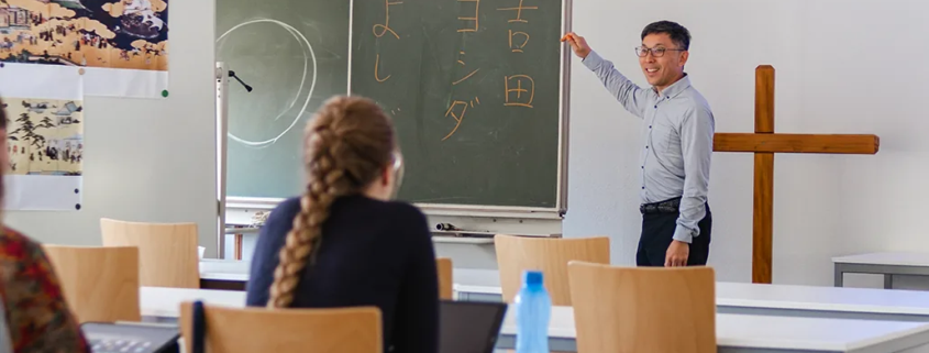 Prof. Dr. Shin Yoshida von der Tohoku Gakuin Universität lehrt während der September School 2026 an der Internationalen Hochschule Liebenzell. Er steht lächelnd vor einer Wandtafel mit japanischen Schriftzeichen und zeigt auf die Tafel, während Studierende im Seminarraum zuhören.