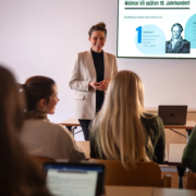 Prof. Dr. Kathrin Thiel steht lächelnd im beigefarbenen Blazer vor einer Präsentationsfolie zum Thema „Weimar im späten 18. Jahrhundert“ und hält eine Vorlesung an der Internationalen Hochschule Liebenzell. Im Vordergrund sind Studierende im Hörsaal zu sehen.