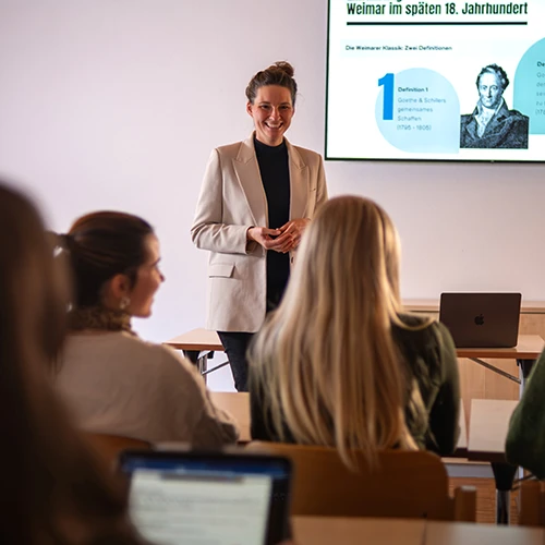 Prof. Dr. Kathrin Thiel steht lächelnd im beigefarbenen Blazer vor einer Präsentationsfolie zum Thema „Weimar im späten 18. Jahrhundert“ und hält eine Vorlesung an der Internationalen Hochschule Liebenzell. Im Vordergrund sind Studierende im Hörsaal zu sehen.