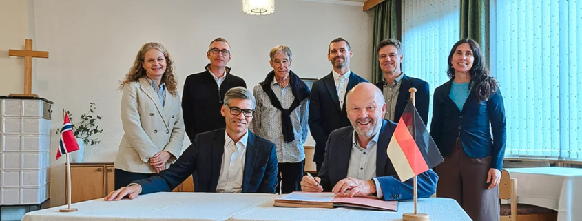 Unterzeichnung der Partnerschaftsvereinbarung (MoU) zwischen der Internationalen Hochschule Liebenzell und dem Fjellhaug International University College. Im Vordergrund sitzen der Rektor des Fjellhaug, Frank-Ole Thoresen, und der IHL-Rektor Volker Gäckle an einem Tisch mit norwegischer und deutscher Tischflagge, während die Delegationsmitglieder beider Hochschulen dahinter stehen.