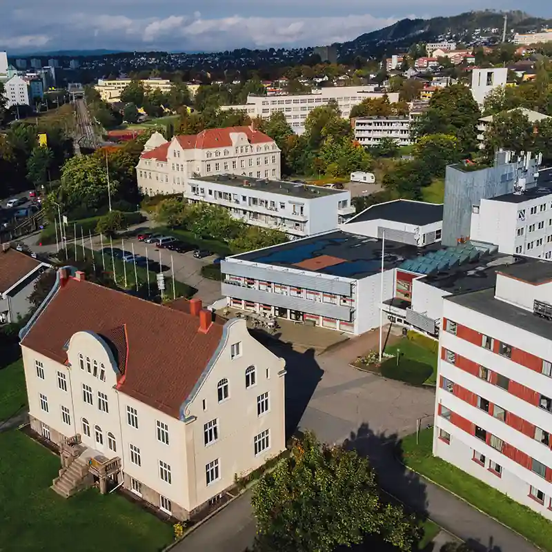 Luftaufnahme des Campus des Fjellhaug International University College (FIUC) in Oslo, einer Partnerhochschule der IHL. Im Vordergrund steht ein historisches Backsteingebäude mit hellem Putz und rotem Ziegeldach, umgeben von modernen Hochschul- und Wohngebäuden vor einer grünen Hügellandschaft.